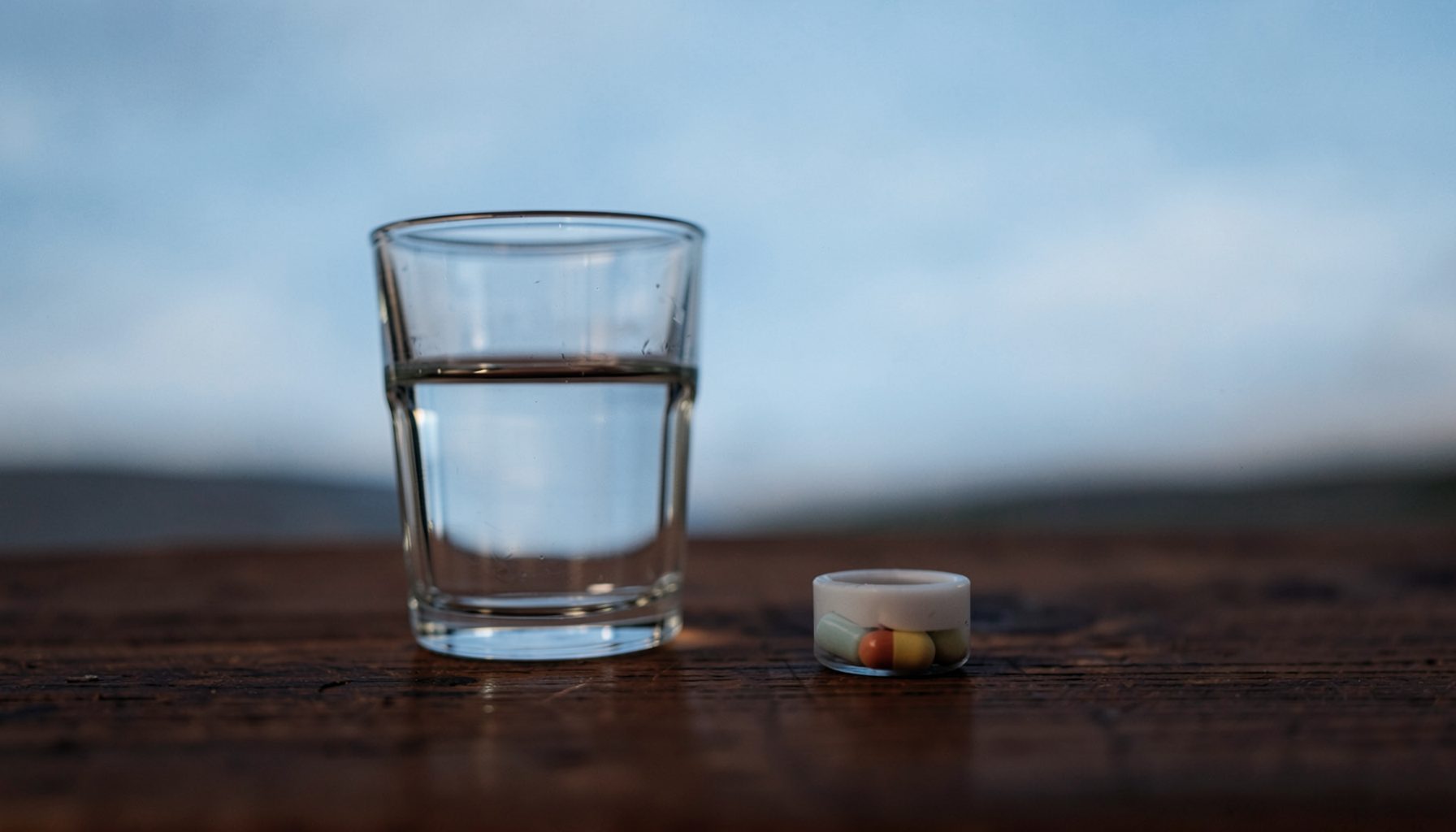 A calm still-life scene in deep brown and soft sky-blue tones, showing a glass of water and a small pill container on a wooden table, evoking a quiet, reassuring mood
