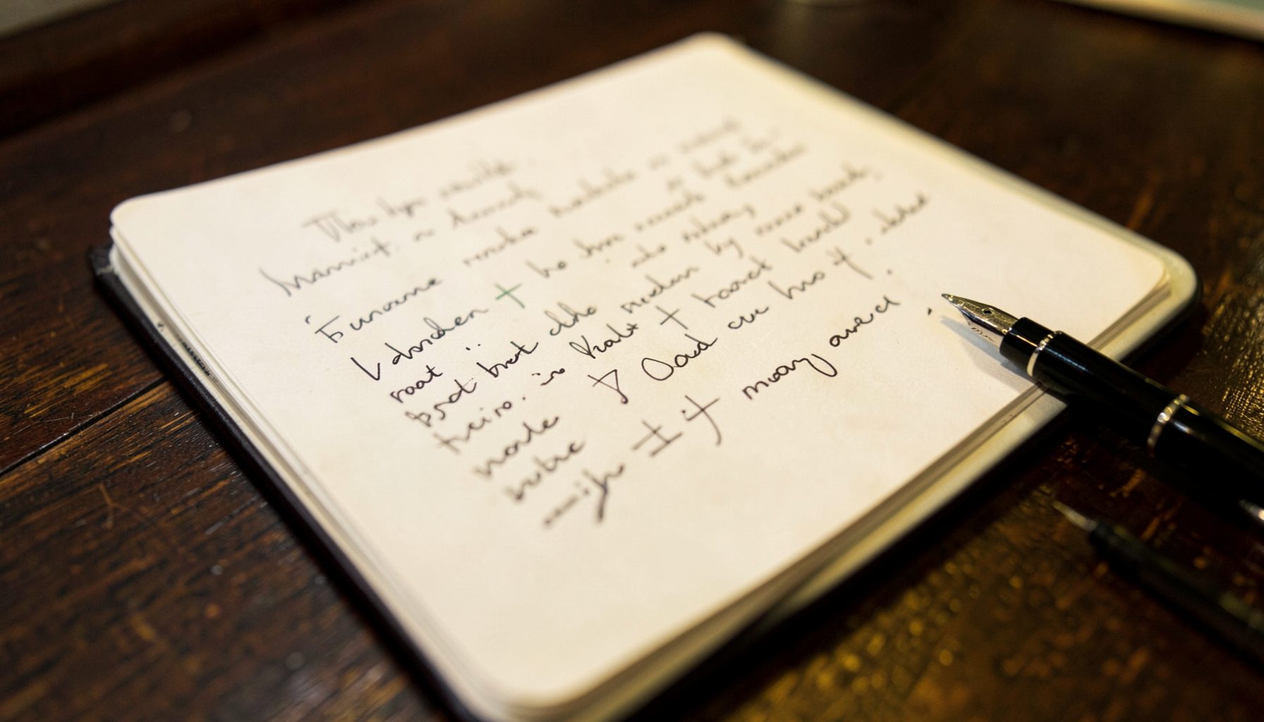 A warm, softly lit close-up of a handwritten journal page with a pen resting beside it, set against a dark wood desk surface in deep brown and muted gold tones.