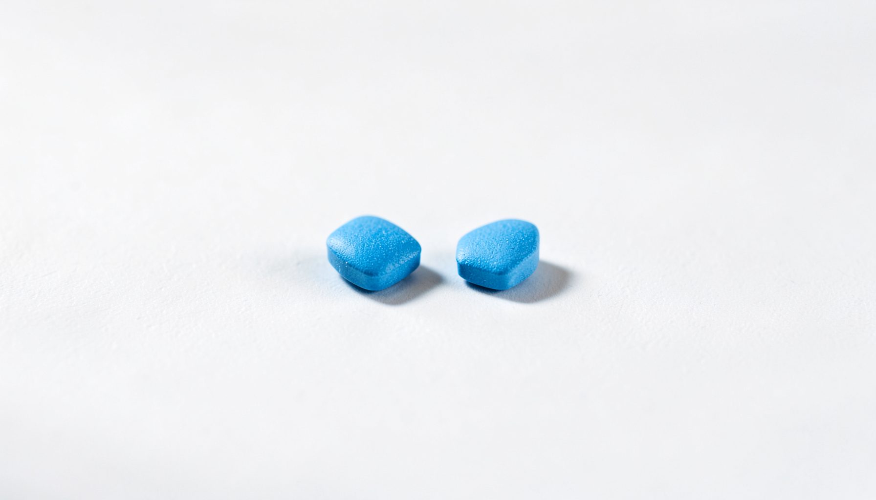 Two small blue diamond-shaped pills resting on a clean white surface, soft natural light casting gentle shadows, calm and clinical mood.